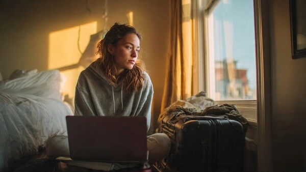 La mentira del nómada digital: lo que nadie te cuenta antes de lanzarte (2026) Mujer sentada con un portátil y una maleta en una habitación luminosa, reflexionando sobre si merece la pena ser nómada digital