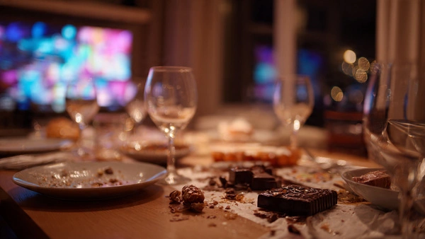 Mesa de Navidad desordenada después de la cena, con copas y restos de turrón, iluminada con luz cálida mientras una televisión aparece desenfocada al fondo.